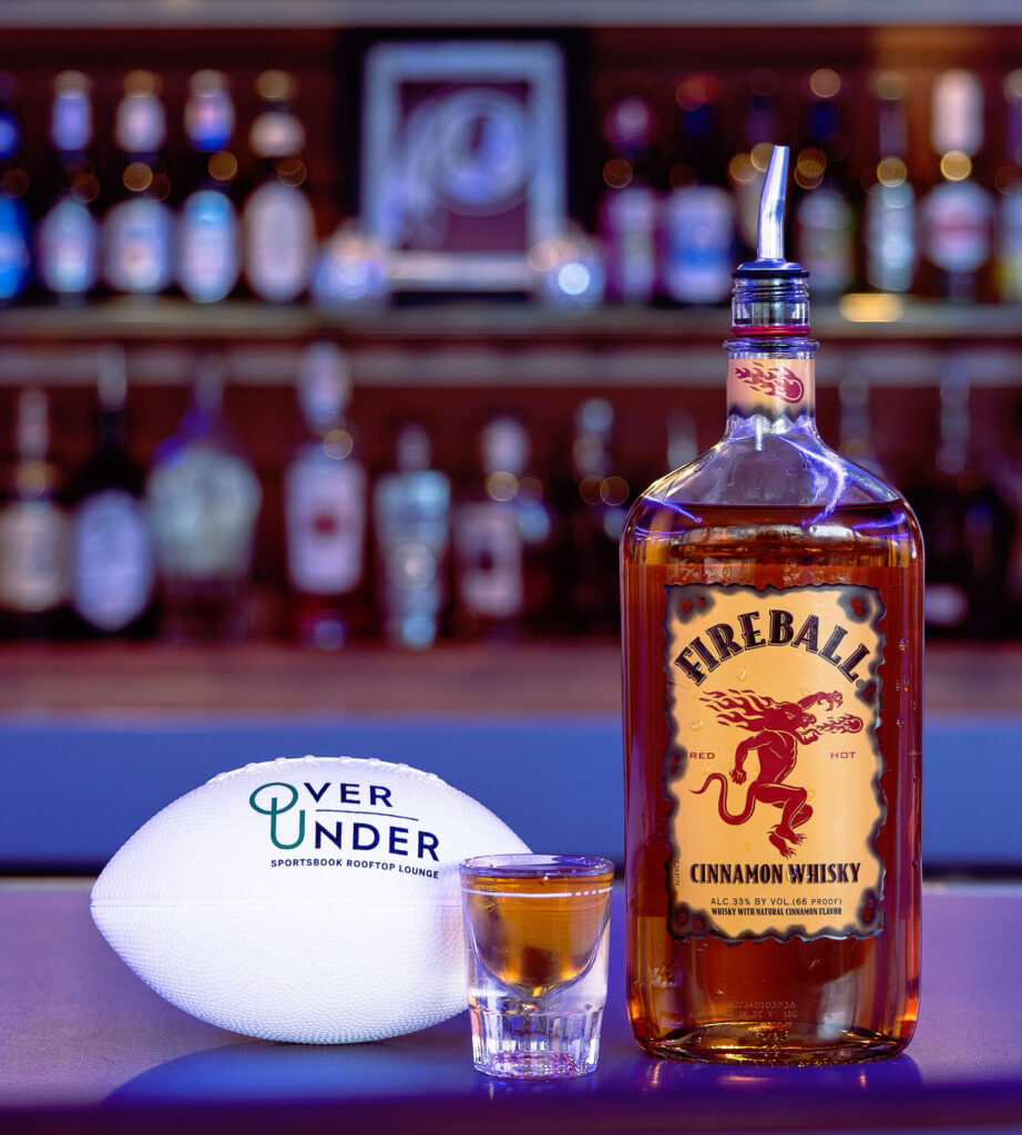 HOME 3 Bottle of Fireball Cinnamon Whisky with a full shot glass beside it and a white Over Under football on a bar counter, with colorful liquor bottles blurred in the background.