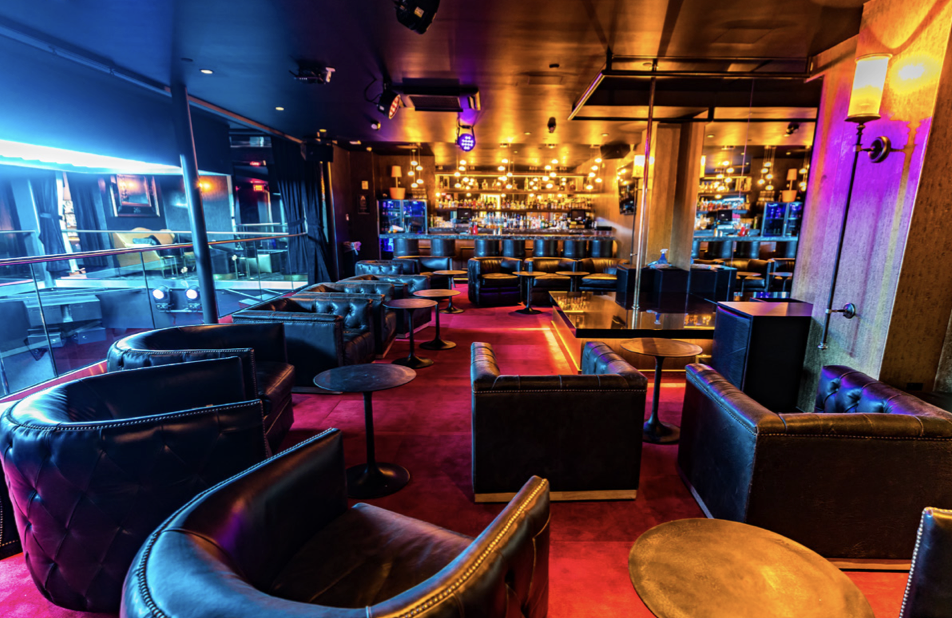 Modern VIP lounge at Cloakroom featuring black leather seating, red carpet flooring, small round tables, and a glowing bar area with warm ambient lighting in the background.