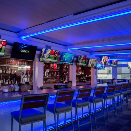 A modern bar area illuminated with bright blue LED lighting, featuring a long counter lined with high bar stools and multiple TV screens mounted above the bar.