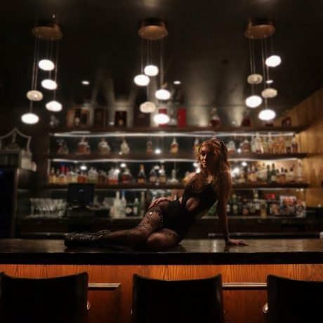 Woman in a black bodysuit and fishnet tights posing on top of a bar counter, with warm hanging lights above and a fully stocked back bar glowing in the background.