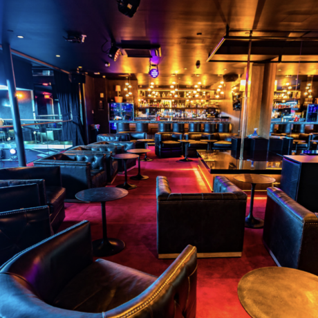 Modern VIP lounge at Cloakroom featuring black leather seating, red carpet flooring, small round tables, and a glowing bar area with warm ambient lighting in the background.